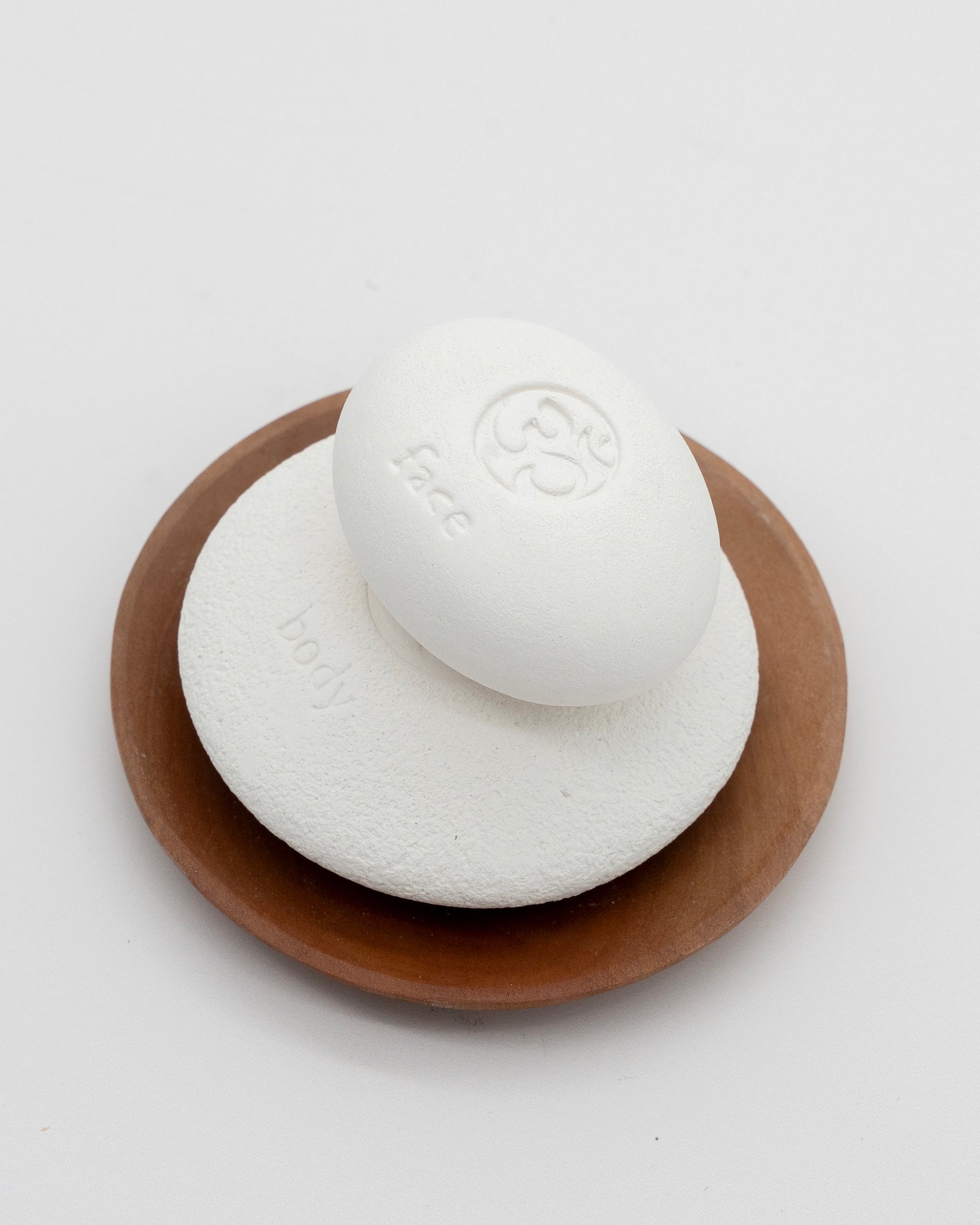 Face Pebble pumice stone in white placed on a round wooden dish, designed for gentle exfoliation and myofascial massage with a smooth, eco-friendly finish.