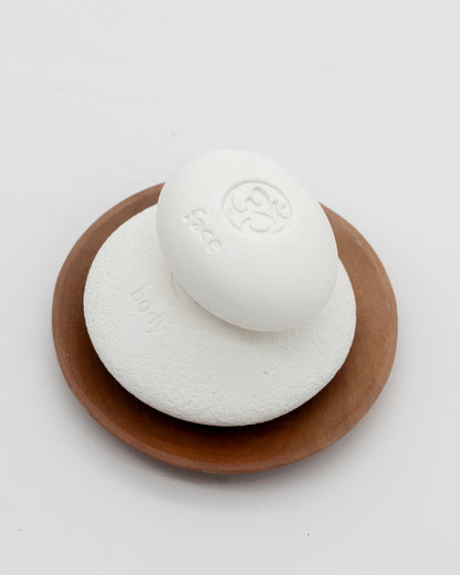 Face Pebble pumice stone in white placed on a round wooden dish, designed for gentle exfoliation and myofascial massage with a smooth, eco-friendly finish.