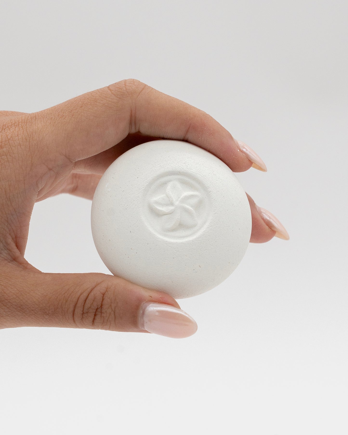 Hand holding a white round Scent Pebble with a subtle embossed flower design, showcasing its smooth texture against a neutral background.