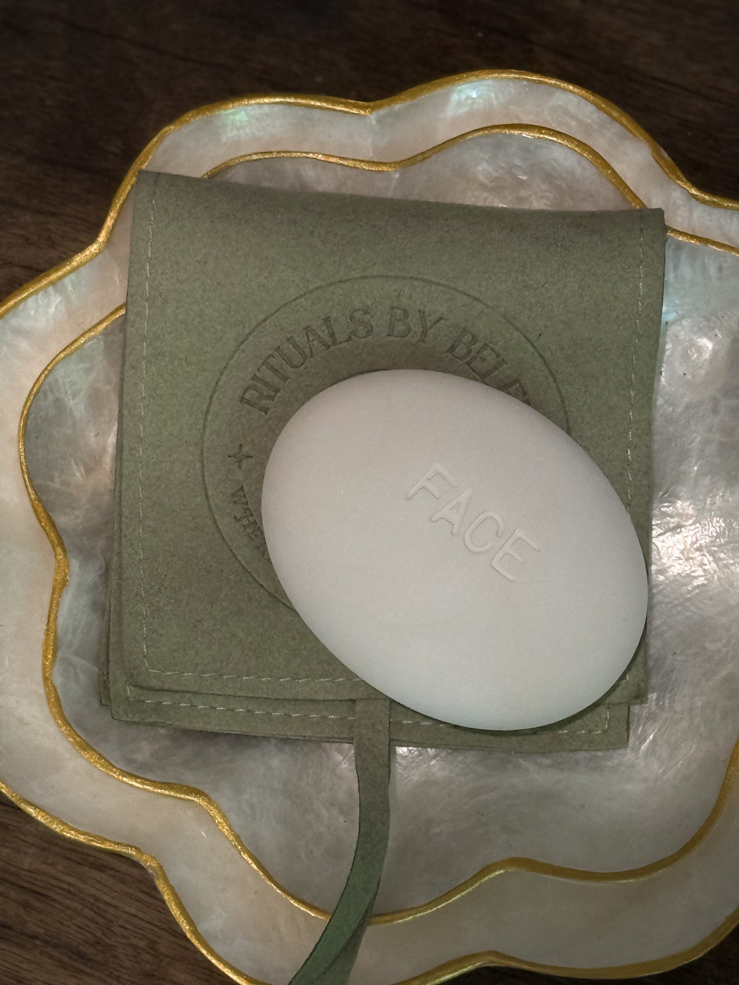 Face Pebble exfoliating stone resting on a green pouch inside a decorative shell-shaped dish with gold trim on a wooden surface