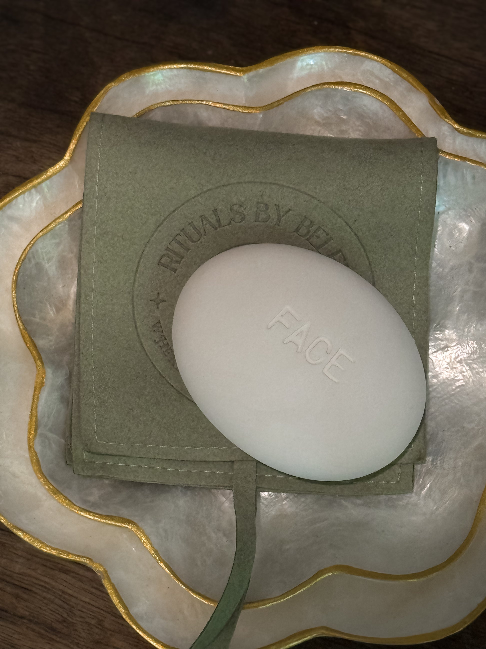 Face Pebble exfoliating stone resting on a green pouch inside a decorative shell-shaped dish with gold trim on a wooden surface
