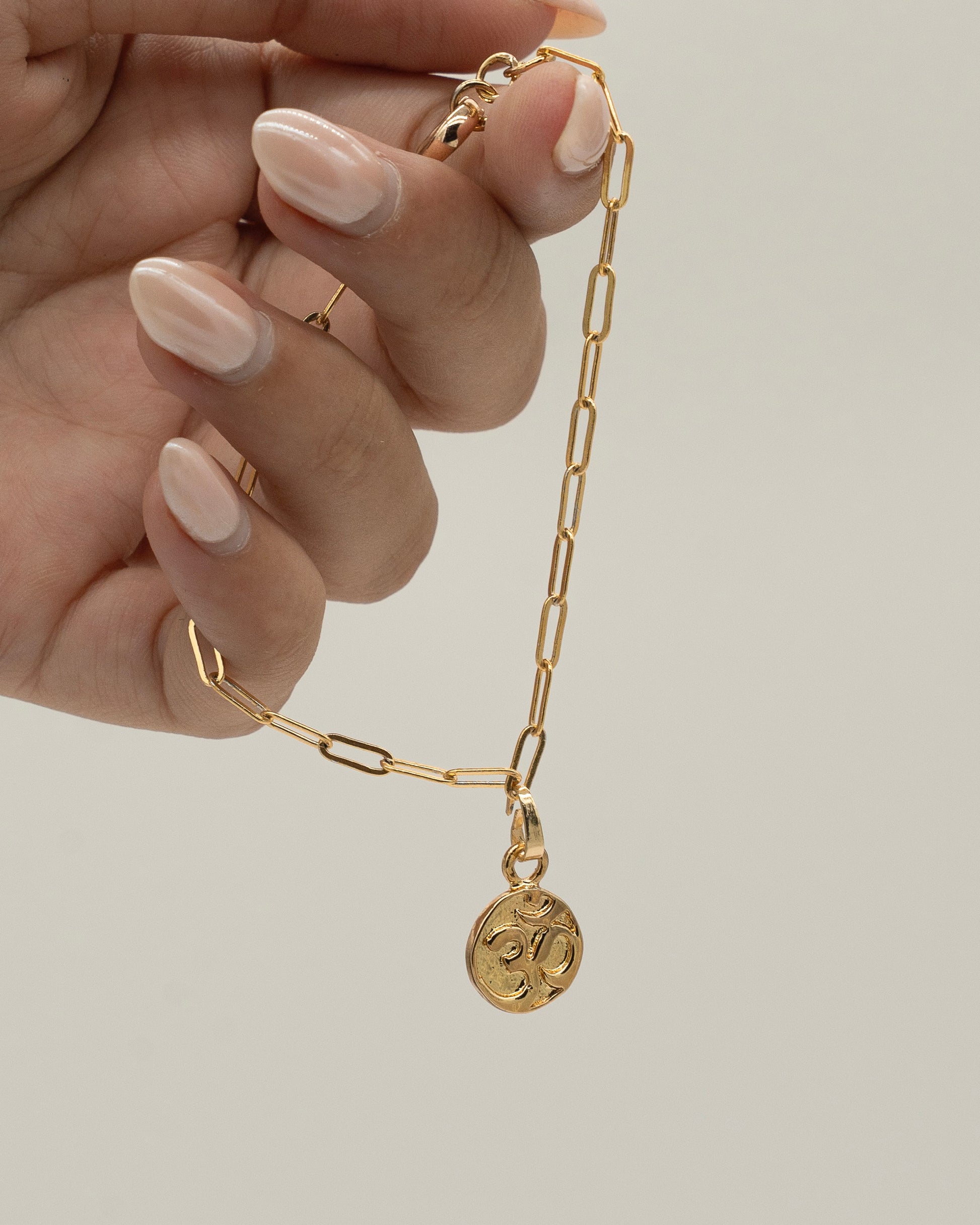 Hand holding a delicate gold Charm Bracelet with rectangular links and a small round charm pendant against a neutral background.