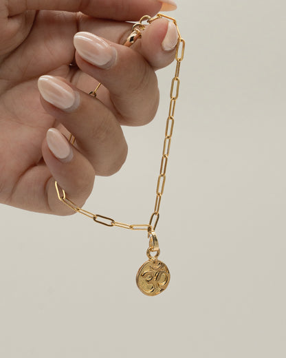 Hand holding a delicate gold Charm Bracelet with rectangular links and a small round charm pendant against a neutral background.