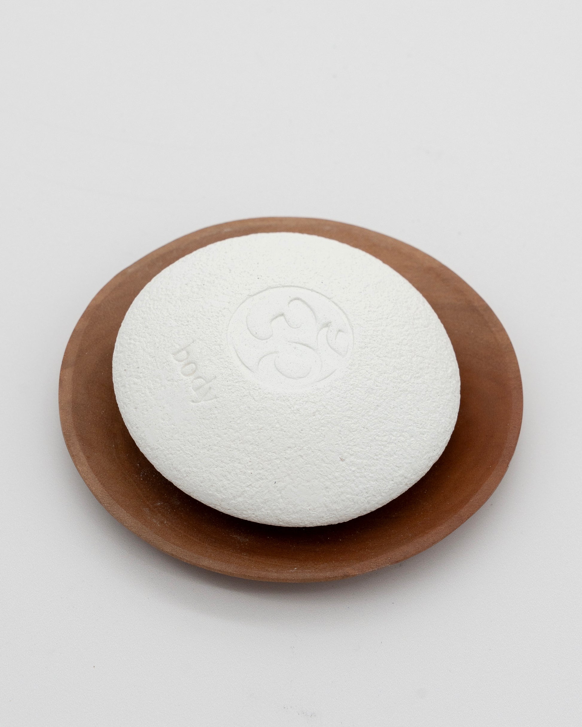White textured Body Pebble pumice stone resting on a brown ceramic dish, designed for exfoliate and myofascial massage.