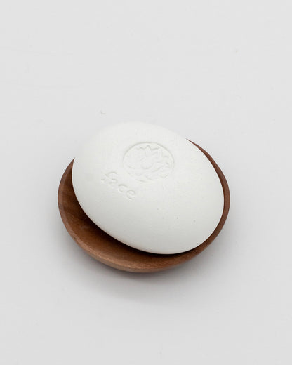 White oval Face Pebble resting on a small round wooden dish, designed for gentle exfoliation and skincare rituals.
