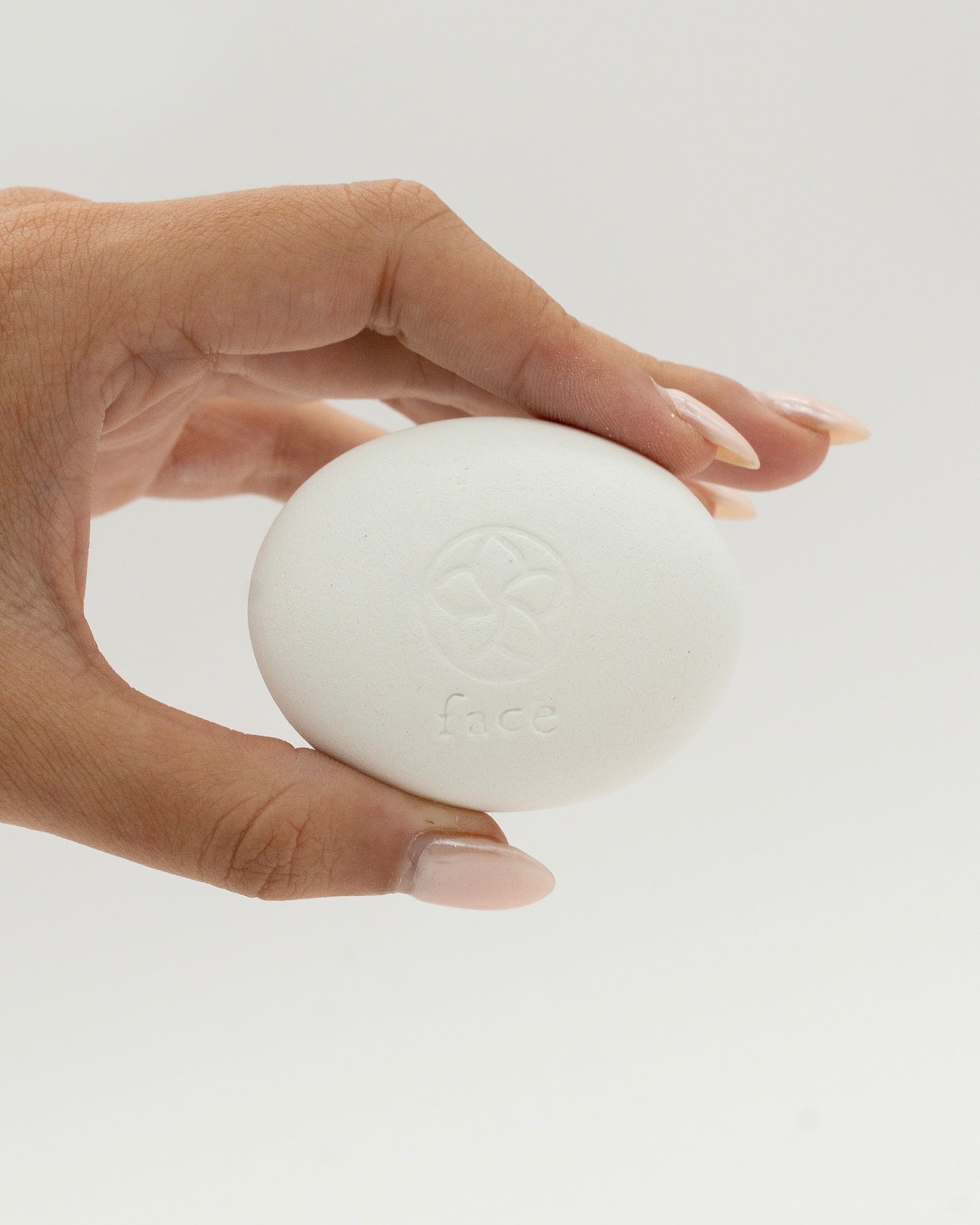 Hand holding a smooth white oval Face Pebble with a subtle embossed design, showcased against a clean, light background for skincare and exfoliation use.