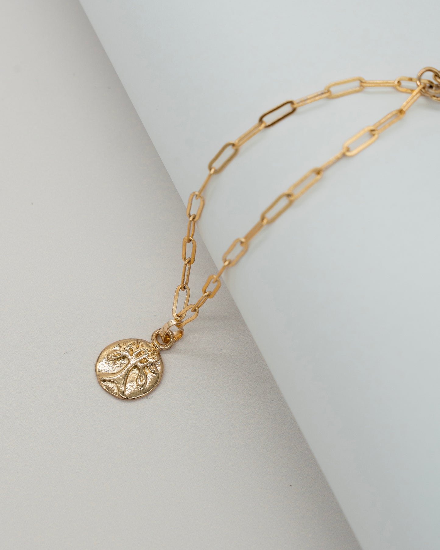 Charm Bracelet featuring a delicate gold chain with elongated links and a small round pendant embossed with an intricate tree design.