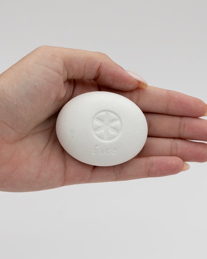 Hand holding a smooth white oval Face Pebble with an embossed circular design and the word face on its surface against a plain light background.