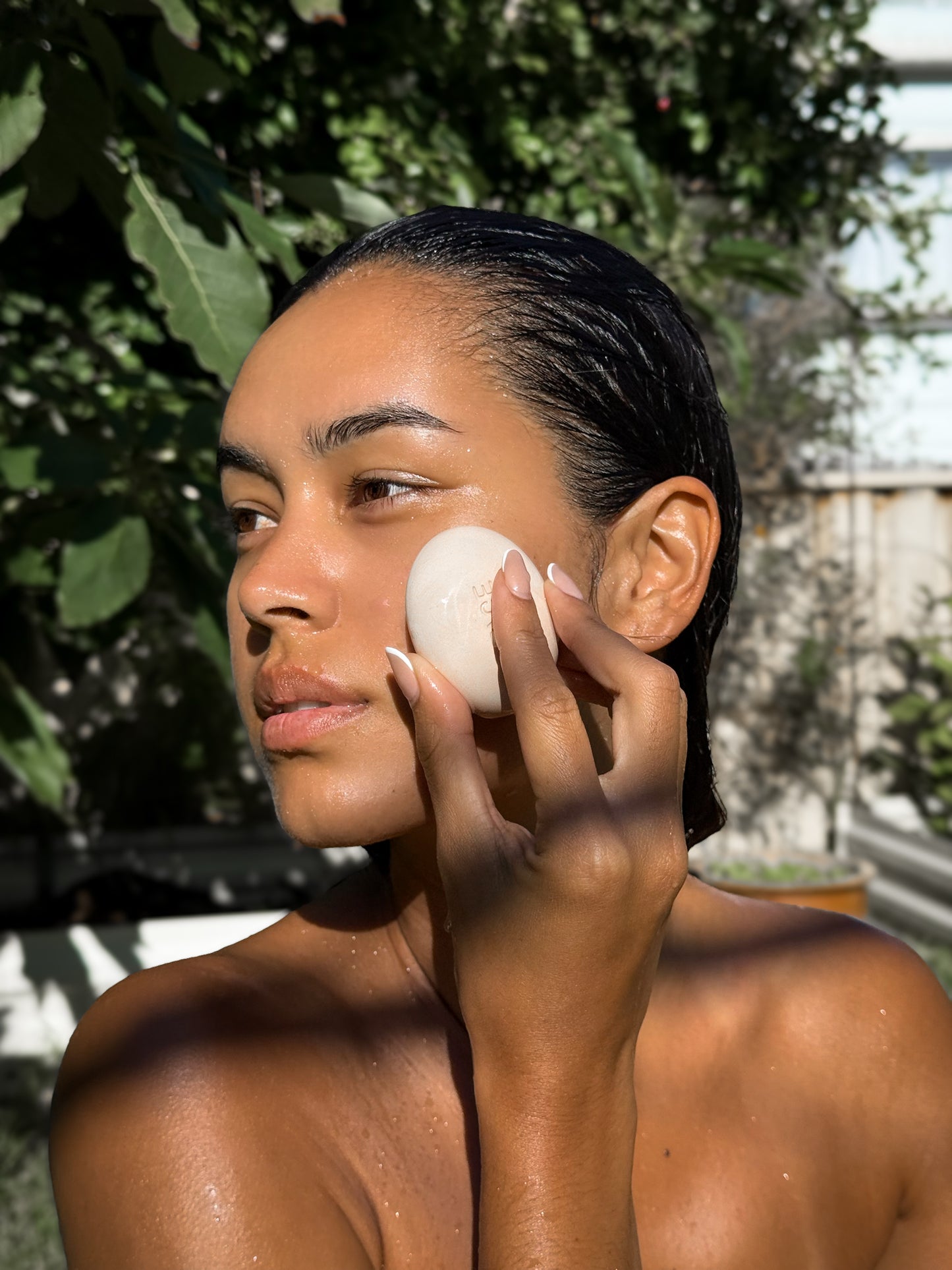 Woman gently applying Face Pebble to her wet cheek in natural outdoor setting, highlighting smooth skin and skincare ritual benefits.