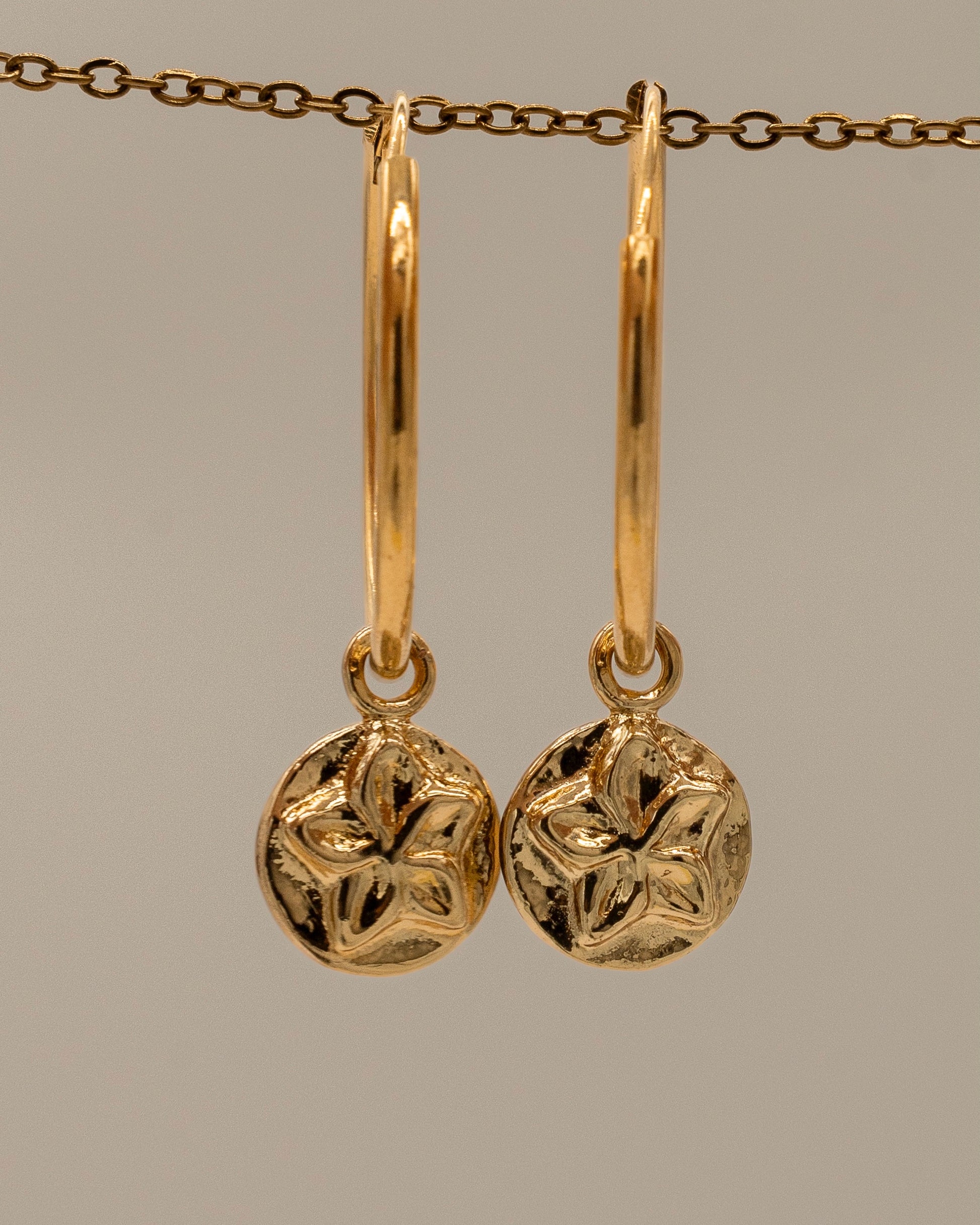 Pair of gold hoop Charm Earrings featuring round floral charm pendants with a textured design hanging from a delicate chain background