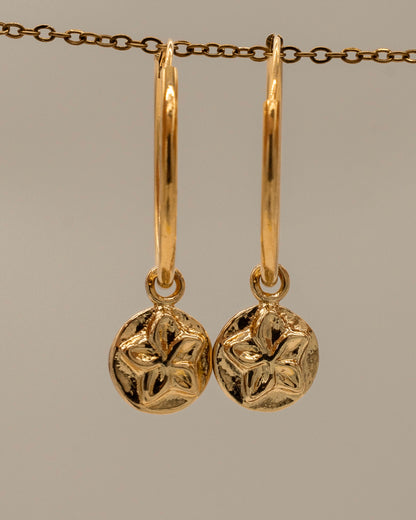 Pair of gold hoop Charm Earrings featuring round floral charm pendants with a textured design hanging from a delicate chain background
