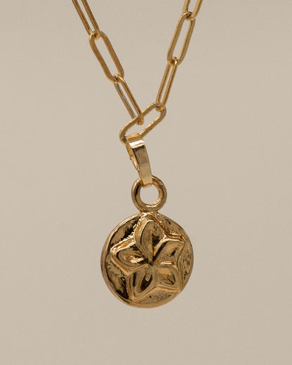 Close-up of a gold Charm Bracelet pendant featuring a textured floral design on a delicate linked chain against a soft beige background.