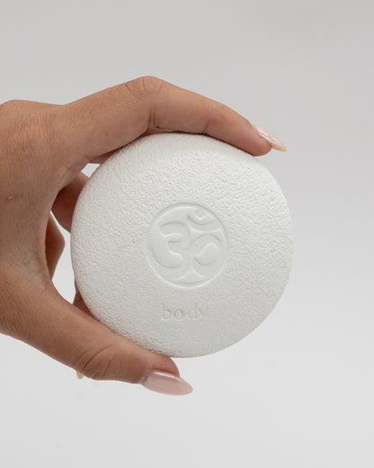 Hand holding a white circular Body Pebble with a textured surface and embossed symbol, designed for exfoliating and myofascial massage.