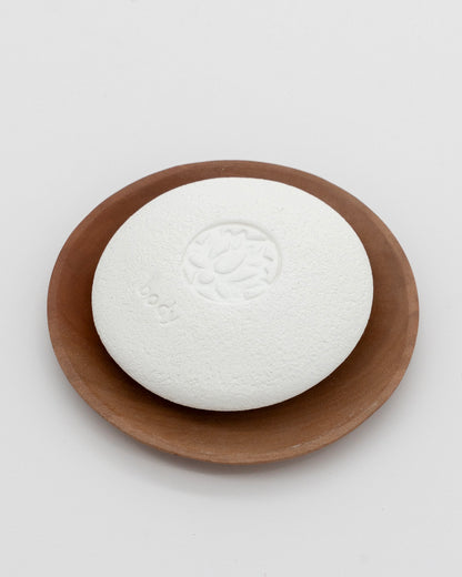 White textured Body Pebble stone with an embossed design, resting on a round wooden dish, ideal for gentle exfoliation and myofascial massage.