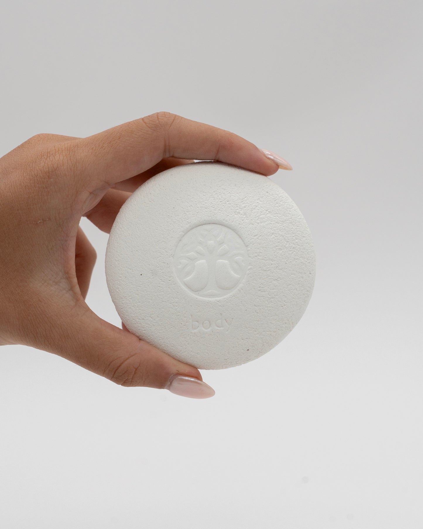 Hand holding a round white textured Body Pebble pumice stone with a subtle embossed design on a plain light background, ideal for exfoliate and myofascial massage.