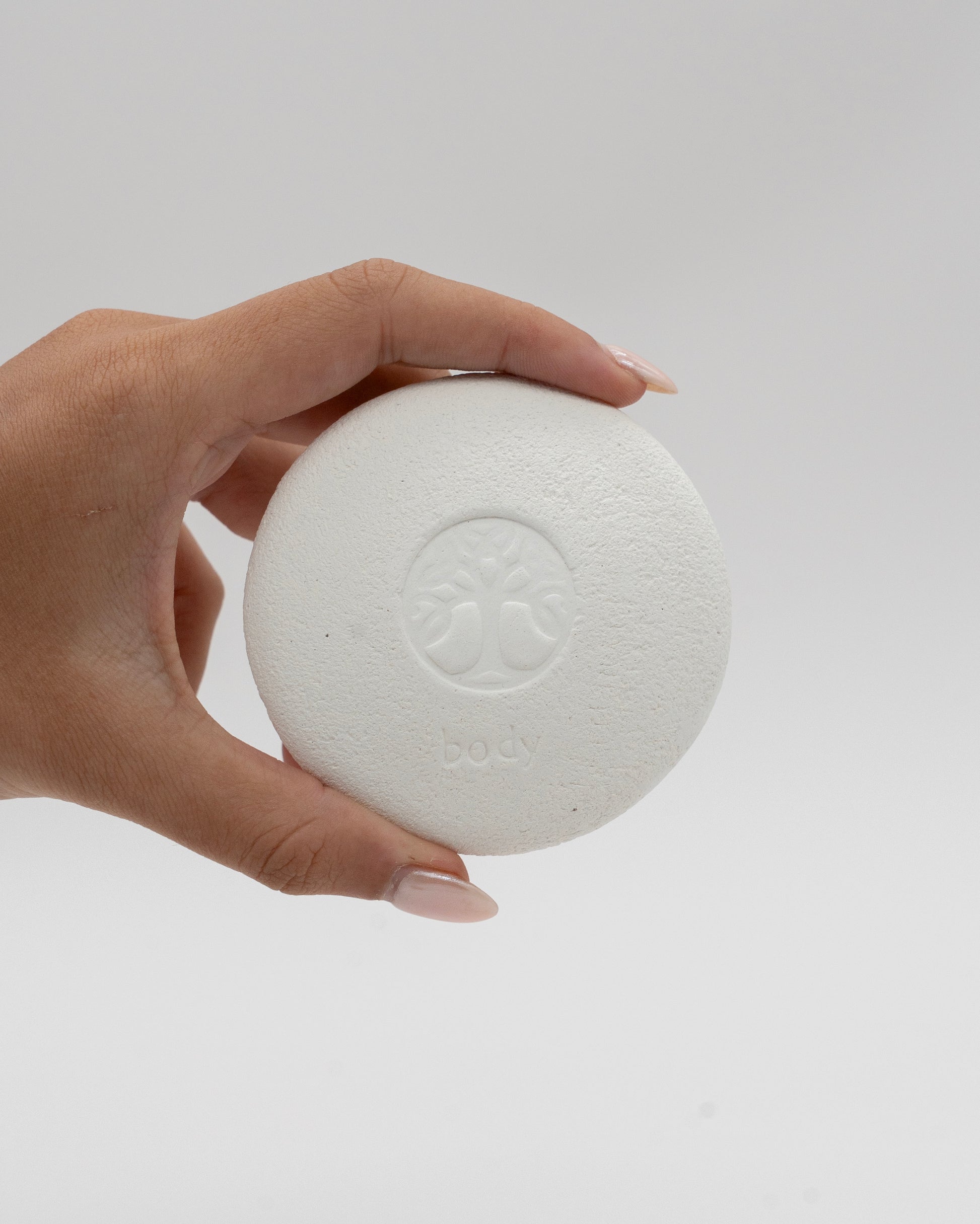 Hand holding a round white textured Body Pebble pumice stone with a subtle embossed design on a plain light background, ideal for exfoliate and myofascial massage.
