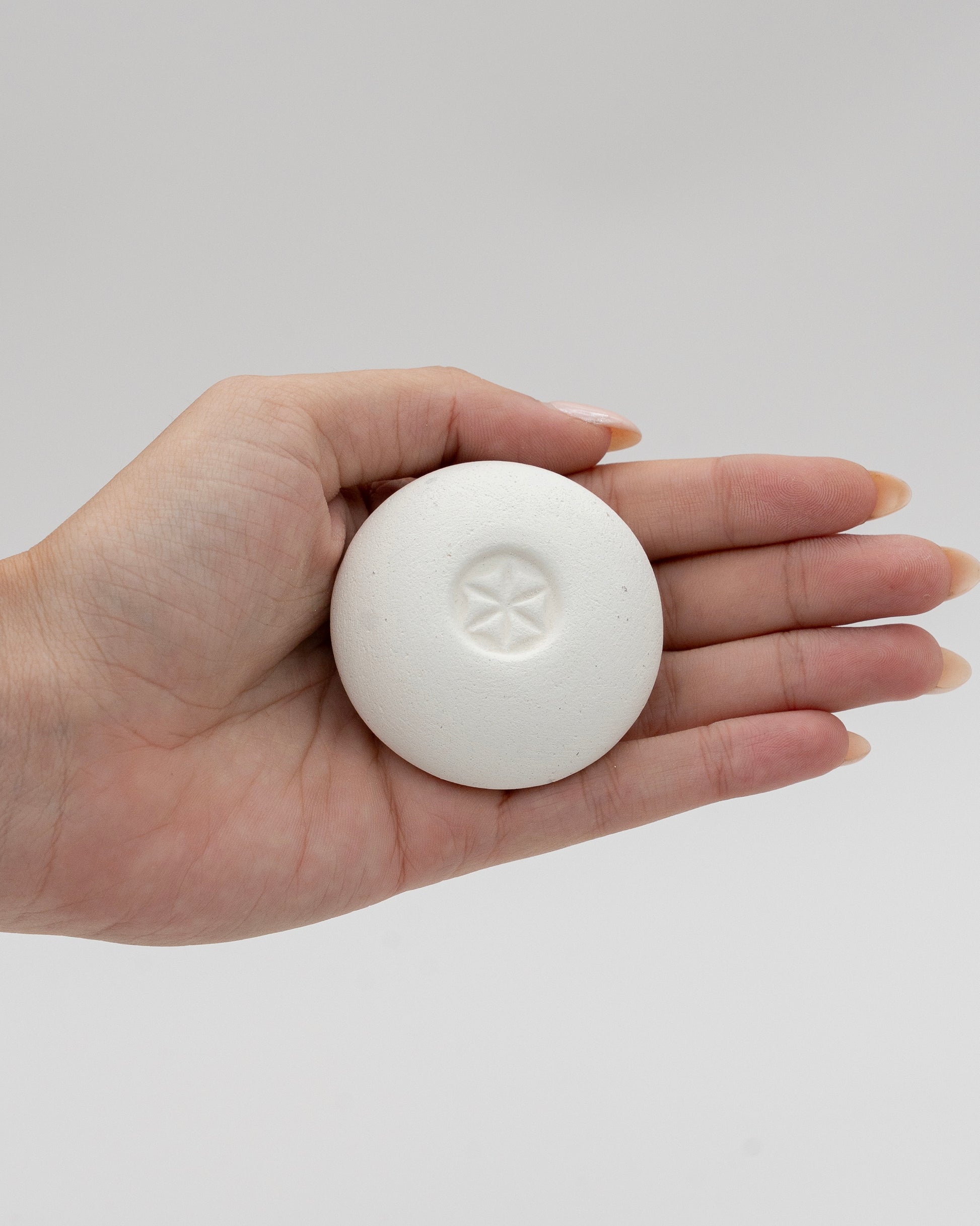 Hand holding a smooth white Scent Pebble with a small embossed star design in the center, showcasing its compact, eco-friendly aromatherapy accessory.