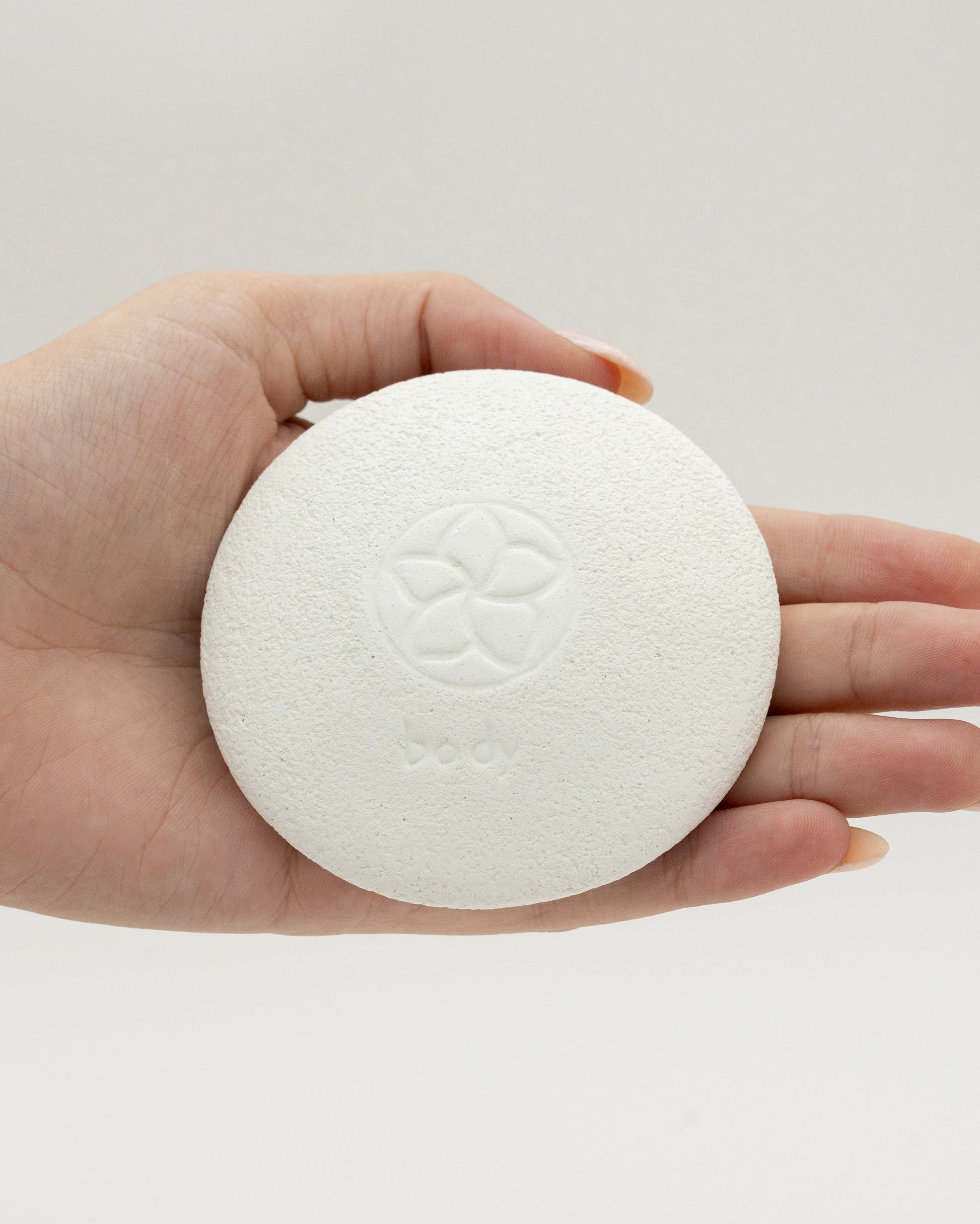 Hand holding a smooth, round white Body Pebble with a subtle embossed floral design, ideal for myofascial massage and exfoliation.