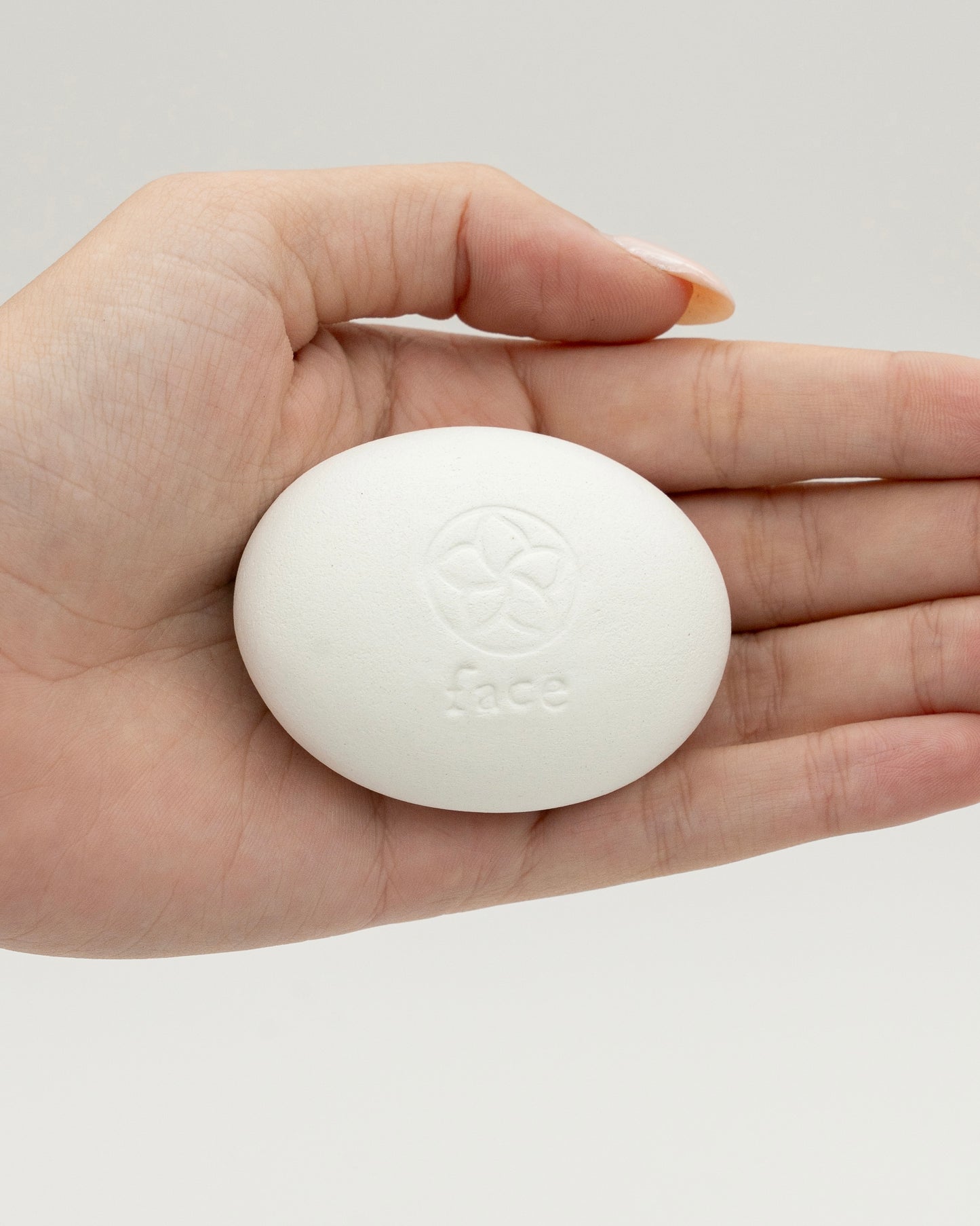 Hand holding a smooth white oval Face Pebble with an embossed floral design and the word "face" on its surface against a plain neutral background.