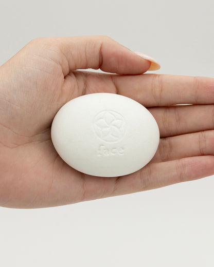 Hand holding a smooth white oval Face Pebble with an embossed floral design and the word "face" on its surface against a plain neutral background.