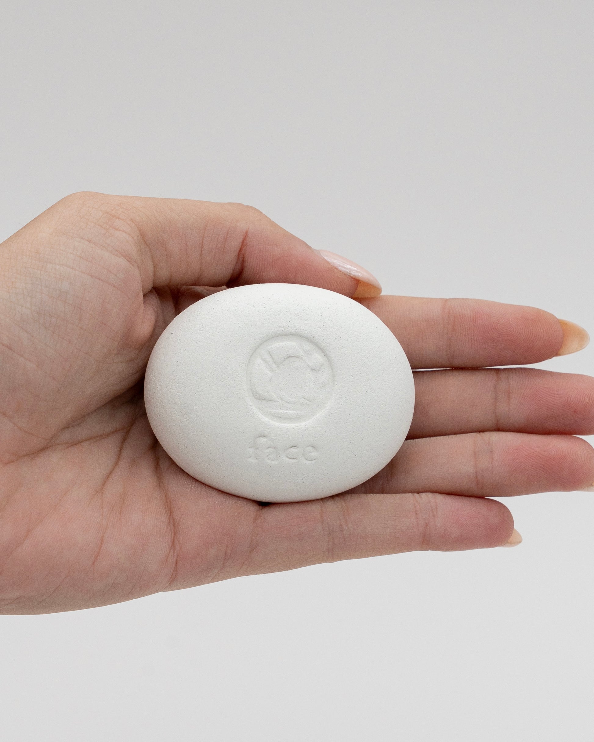 A close-up of a hand holding a smooth white Face Pebble with an embossed logo, designed for gentle exfoliation and myofascial massage.