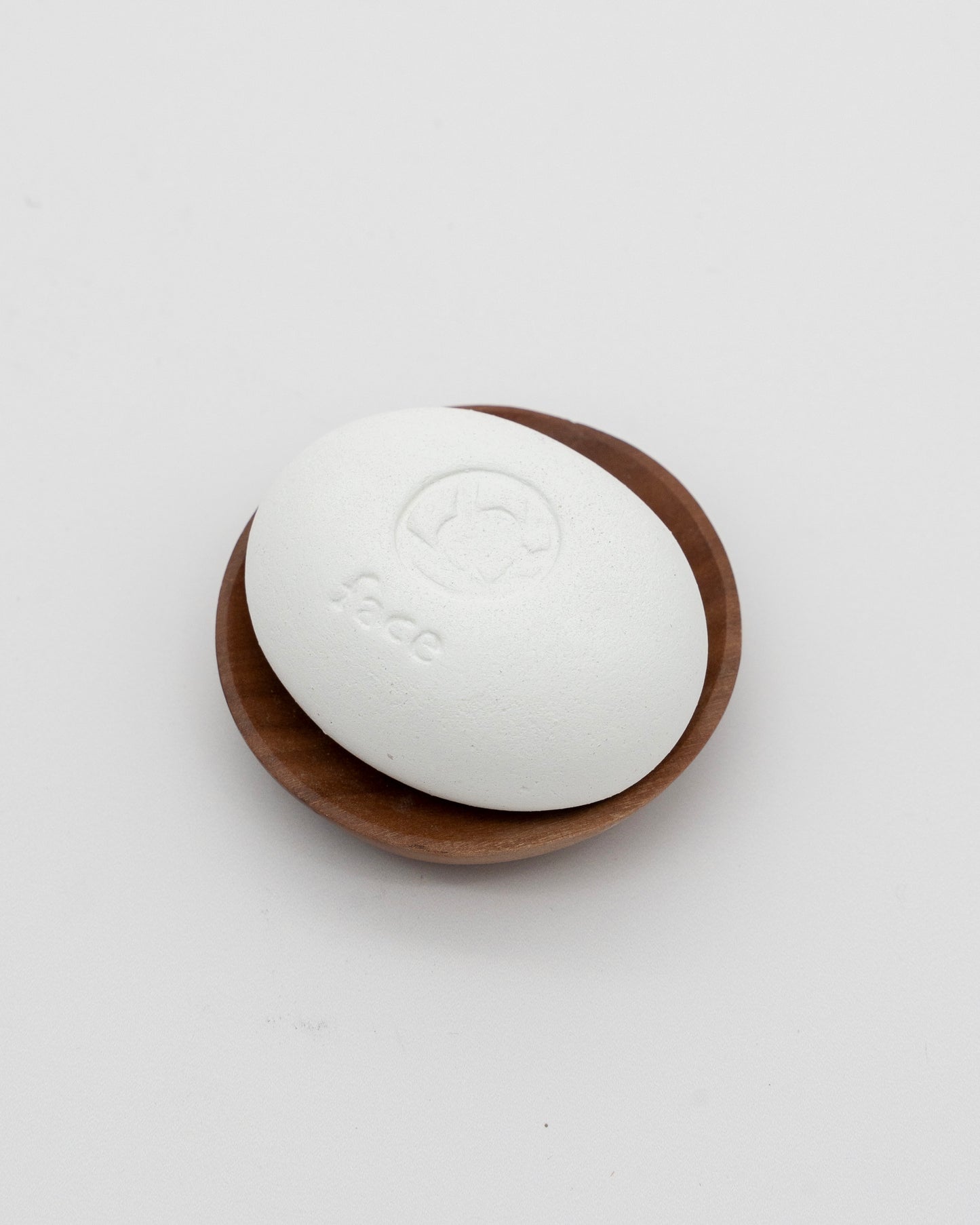 White eco-friendly Face Pebble stone for myofascial massage and exfoliation resting on a small wooden dish against a plain background.
