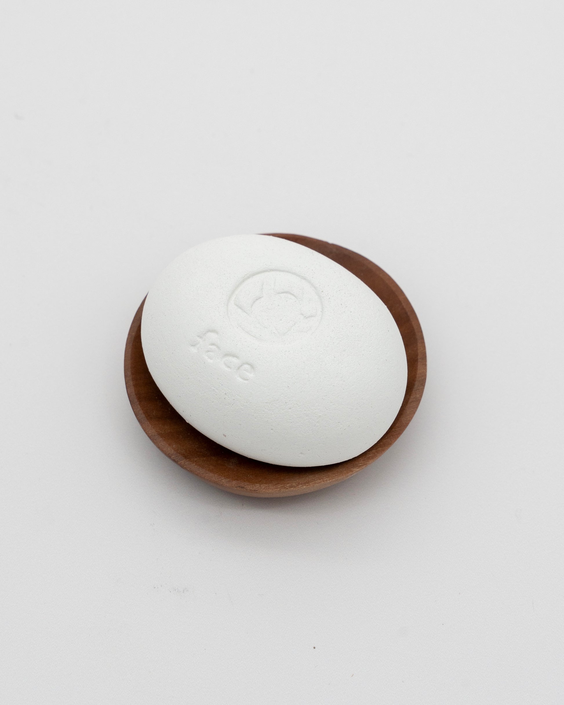 White eco-friendly Face Pebble stone for myofascial massage and exfoliation resting on a small wooden dish against a plain background.