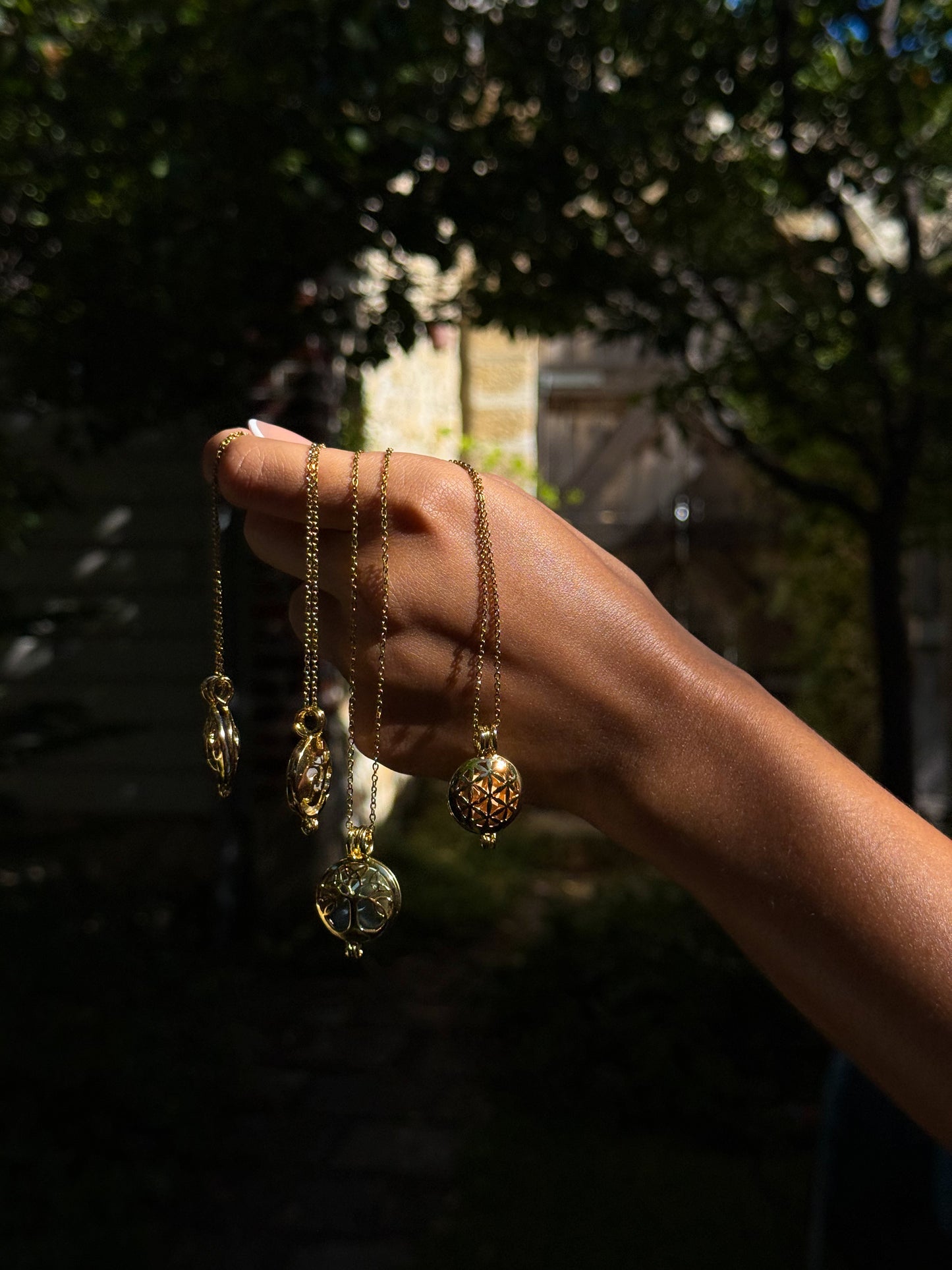 Close-up of a hand holding multiple delicate Gold Scent Locket necklaces with intricate designs, showcased outdoors in natural light against a blurred garden background.