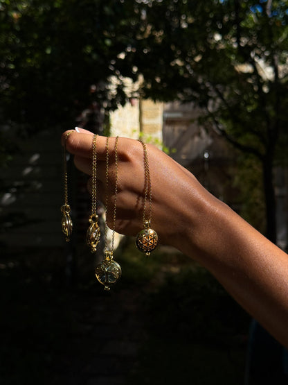 Close-up of a hand holding multiple delicate Gold Scent Locket necklaces with intricate designs, showcased outdoors in natural light against a blurred garden background.