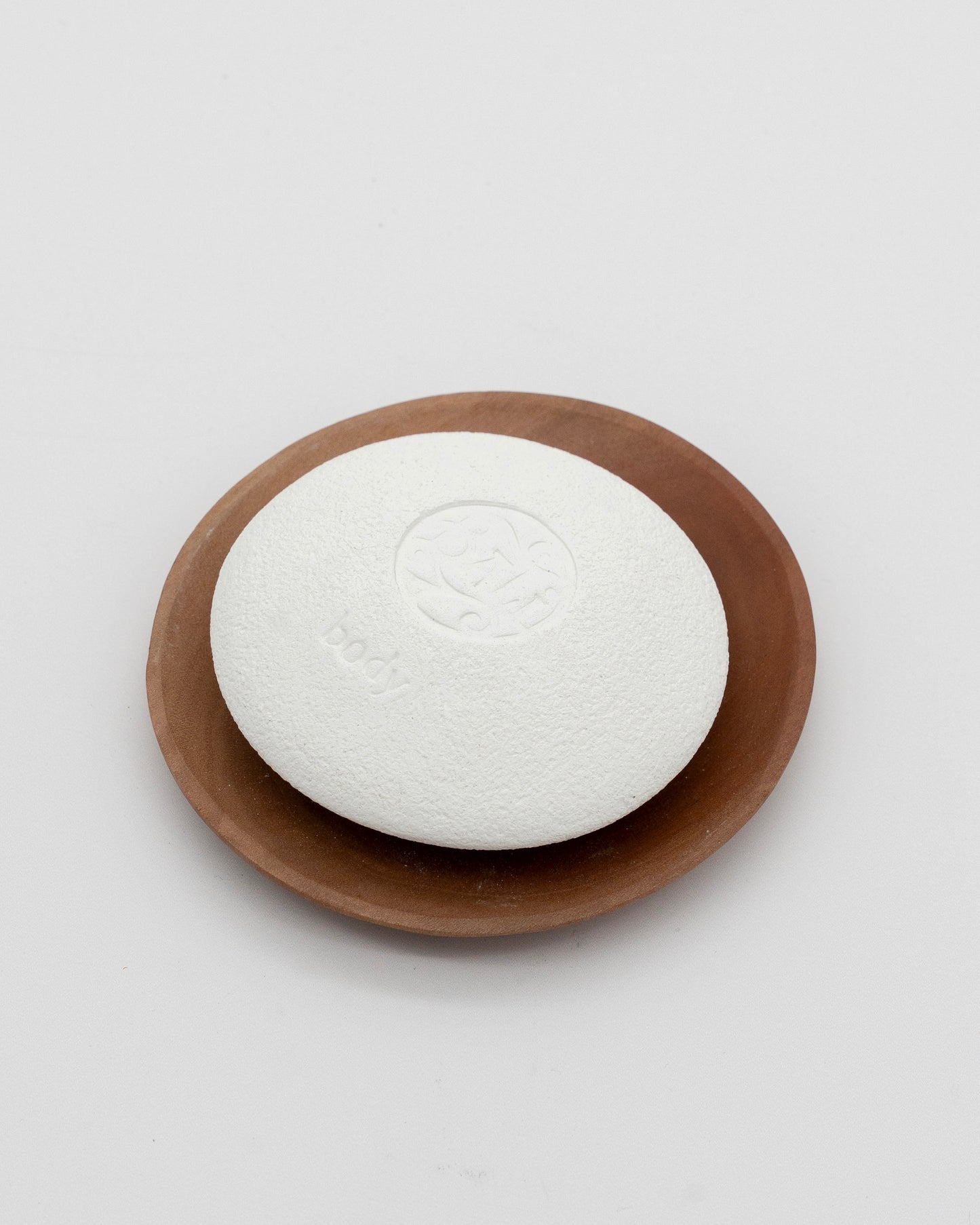 White textured pumice stone with smooth rounded edges labeled Body Pebble, resting on a brown wooden dish against a white background.