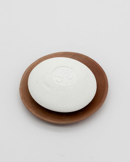 White textured pumice stone with smooth rounded edges labeled Body Pebble, resting on a brown wooden dish against a white background.