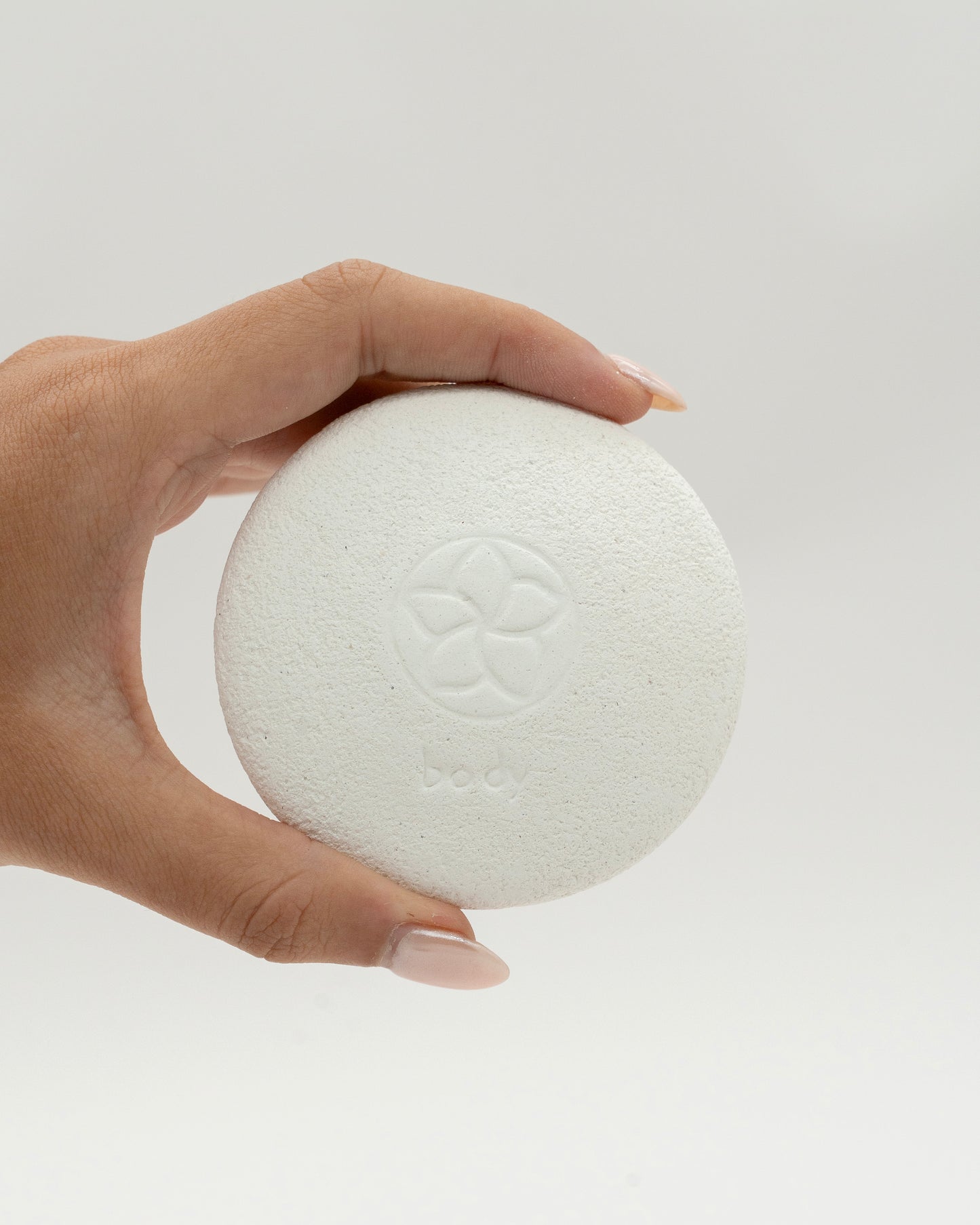 Hand holding a round white Body Pebble pumice stone with a subtle embossed floral design, used for exfoliating and myofascial massage.
