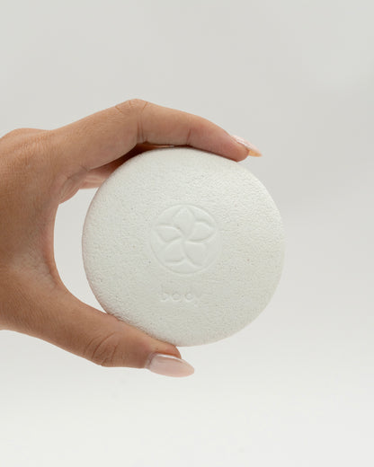 Hand holding a round white Body Pebble pumice stone with a subtle embossed floral design, used for exfoliating and myofascial massage.