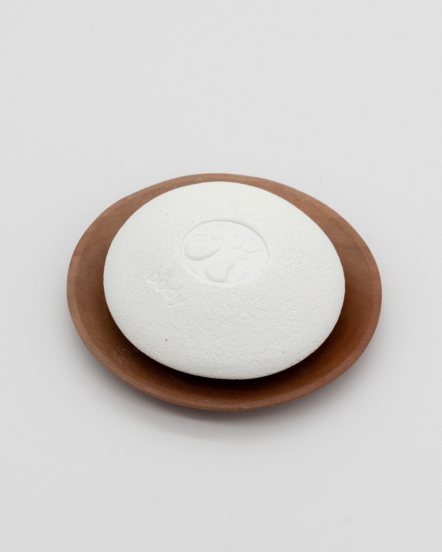 White textured Body Pebble with embossed tree design resting on a smooth brown wooden dish against a plain white background