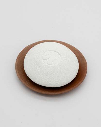 White textured Body Pebble with embossed tree design resting on a smooth brown wooden dish against a plain white background