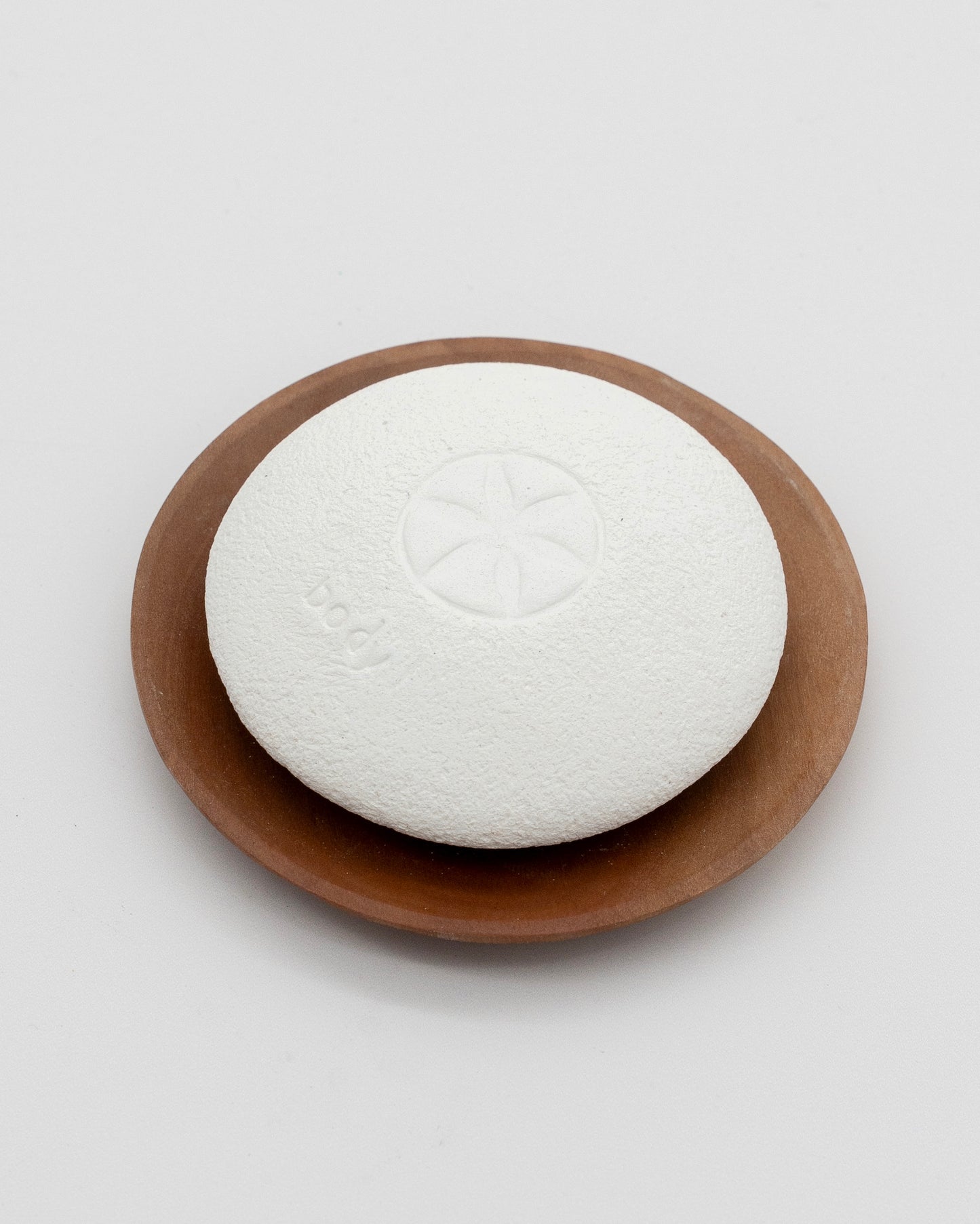 White textured Body Pebble resting on a round brown wooden dish, designed for gentle exfoliation and myofascial massage.