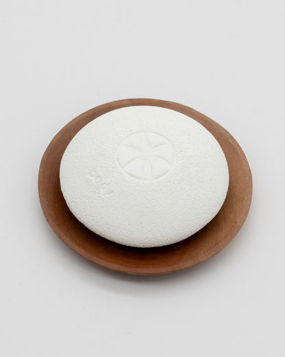 White textured Body Pebble resting on a round brown wooden dish, designed for gentle exfoliation and myofascial massage.