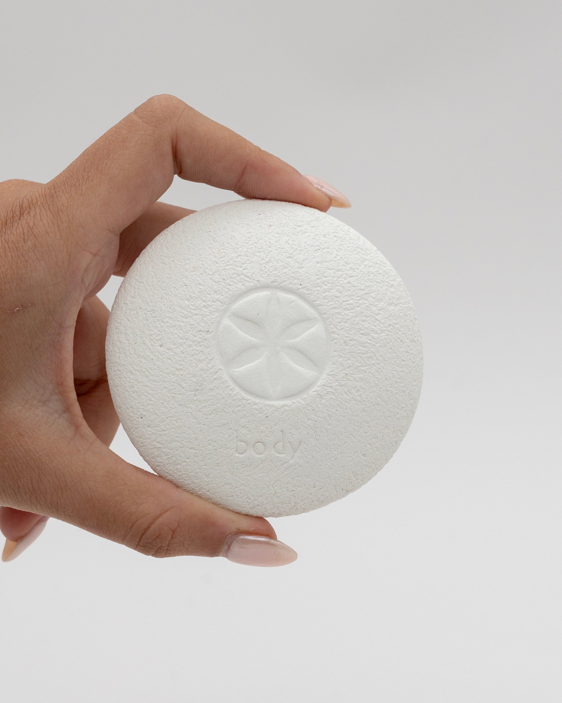 Hand holding a round white textured Body Pebble with a subtle star-shaped design and the word "body" embossed on its surface against a plain background.