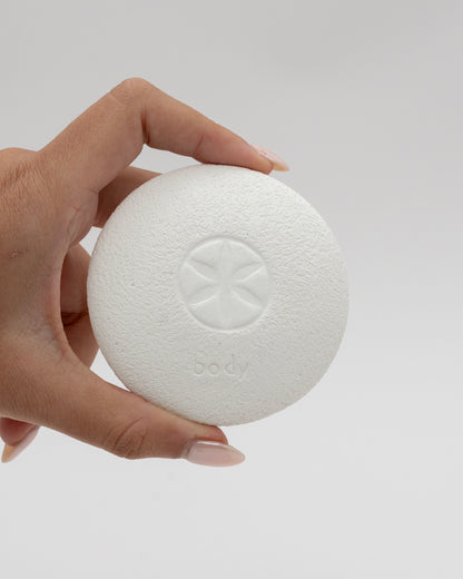Hand holding a round white textured Body Pebble with a subtle star-shaped design and the word "body" embossed on its surface against a plain background.