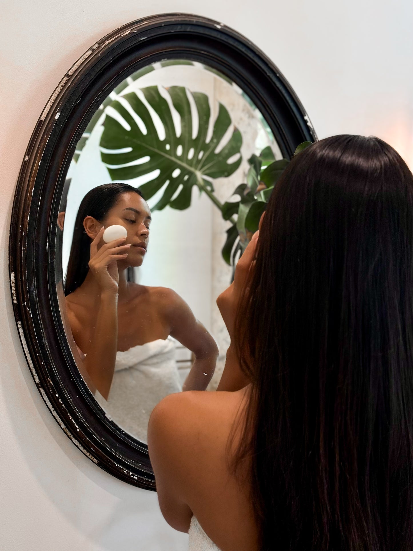 Woman with long dark hair wrapped in a towel using a Face Pebble for gentle exfoliation and myofascial massage in front of a round black-framed mirror with green plant reflection.