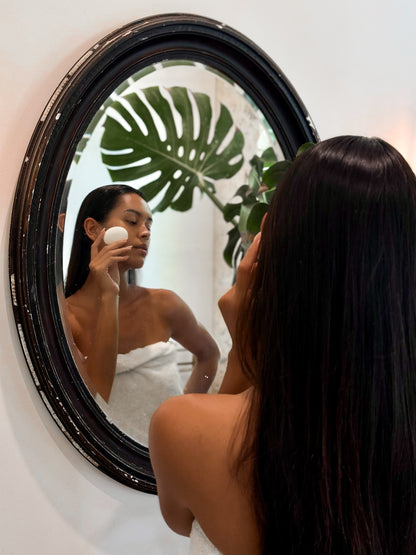 Woman with long dark hair wrapped in a towel using a Face Pebble for gentle exfoliation and myofascial massage in front of a round black-framed mirror with green plant reflection.