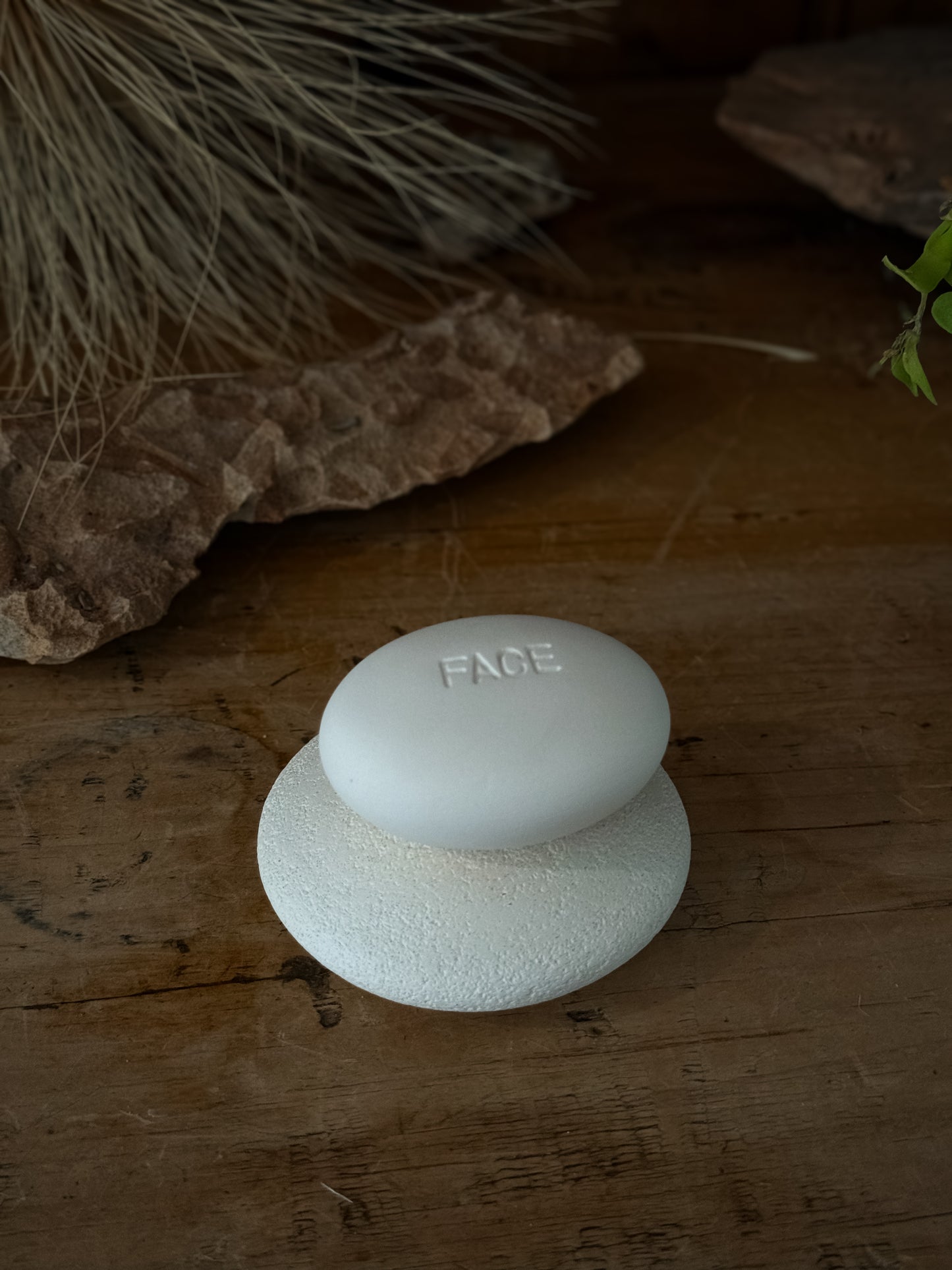 Minimalist white Face Pebble resting on a textured round base on a wooden surface, with natural dried grass and rustic elements in the background for an eco friendly aesthetic.