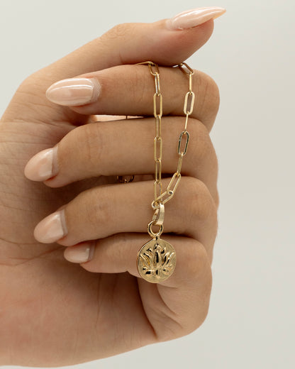 Hand holding a delicate gold Charm Bracelet with elongated chain links and a round lotus flower charm pendant against a neutral background.