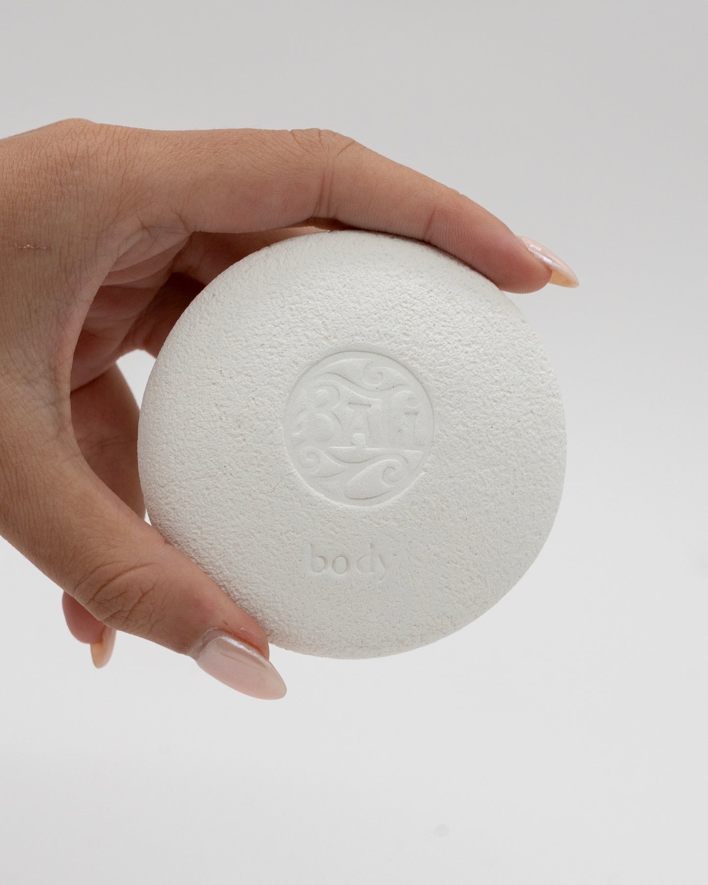 Hand holding a white textured circular Body Pebble with an embossed logo and the word body, designed for exfoliation and myofascial massage.