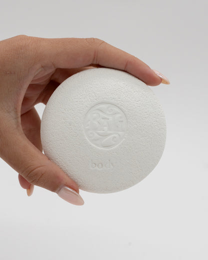 Hand holding a white textured circular Body Pebble with an embossed logo and the word body, designed for exfoliation and myofascial massage.