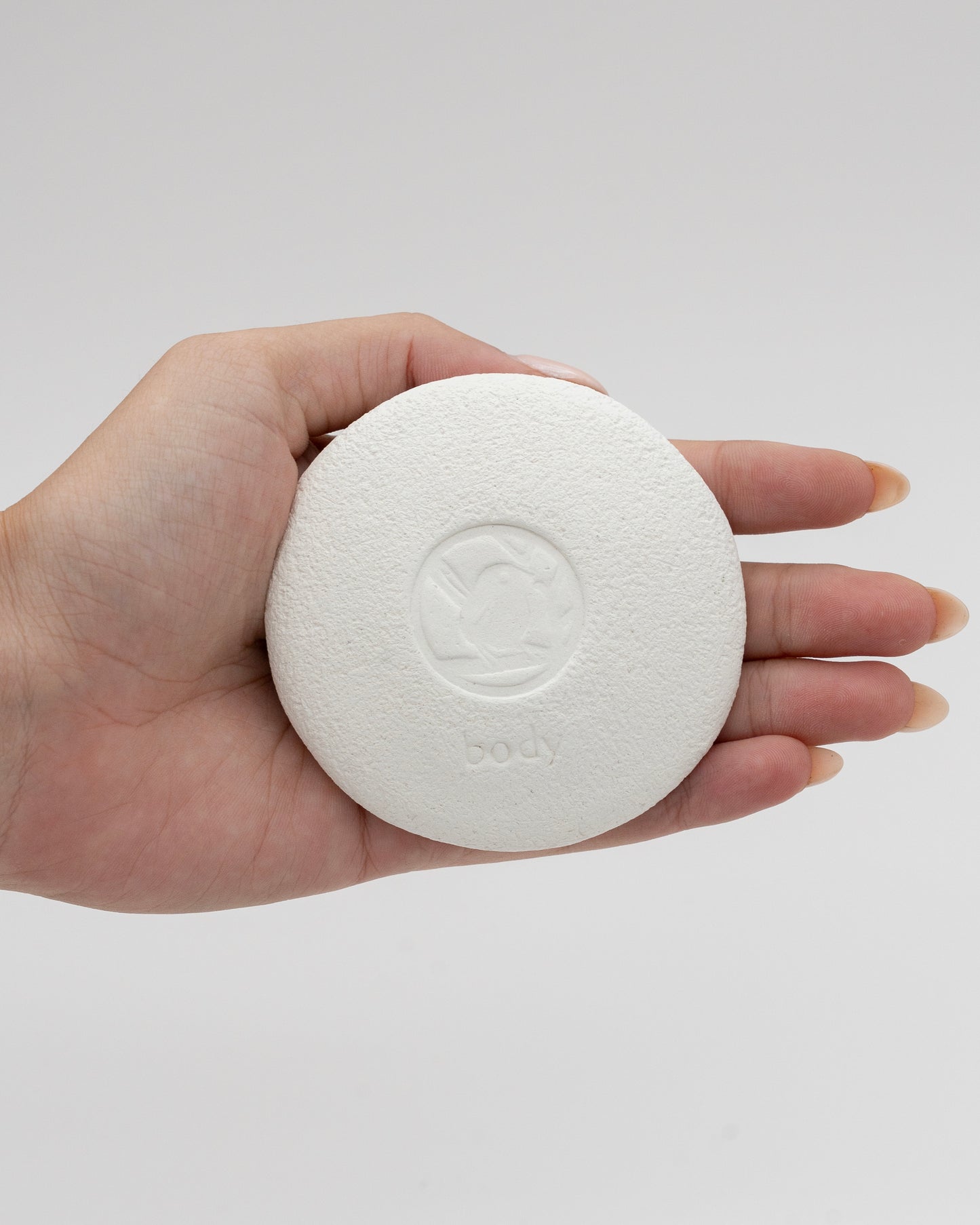 Hand holding a white textured circular Body Pebble, designed for myofascial massage and exfoliation, with a subtle logo embossed in the center.