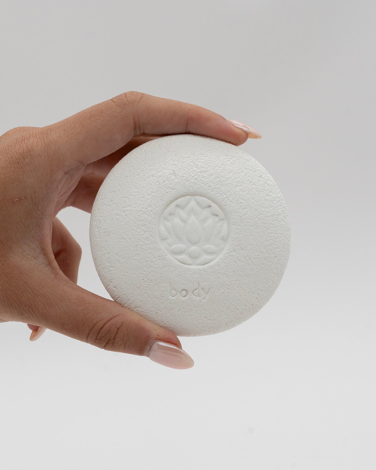 Hand holding a white circular Body Pebble with a lotus flower design embossed in the center, used for myofascial massage and exfoliating skin gently.