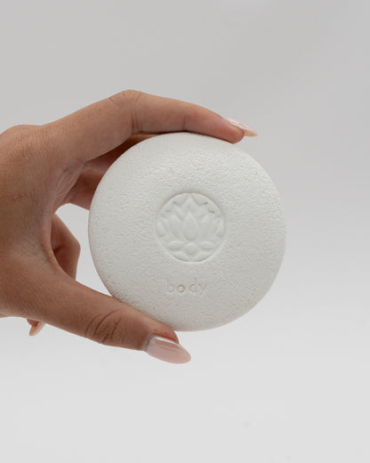Hand holding a white circular Body Pebble with a lotus flower design embossed in the center, used for myofascial massage and exfoliating skin gently.