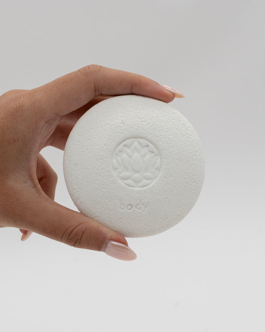 Hand holding a white circular Body Pebble with a lotus flower design embossed in the center, used for myofascial massage and exfoliating skin gently.