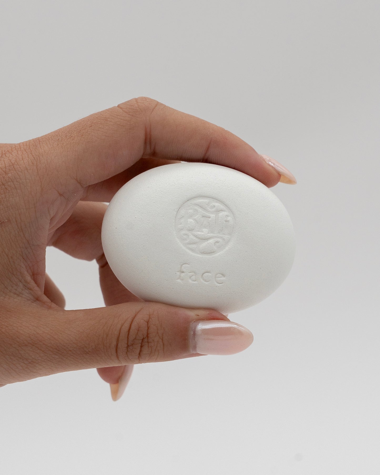 Hand holding a smooth white face stone with a subtle embossed design and the word Face Pebble, ideal for gentle exfoliation and myofascial massage.